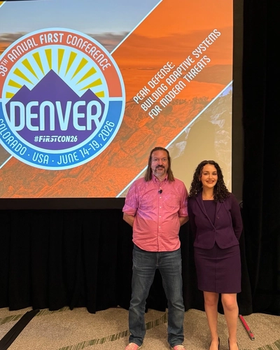 Rina Rakipi and Tom Millar stand smiling in front of a FIRST Conference screen in Denver.