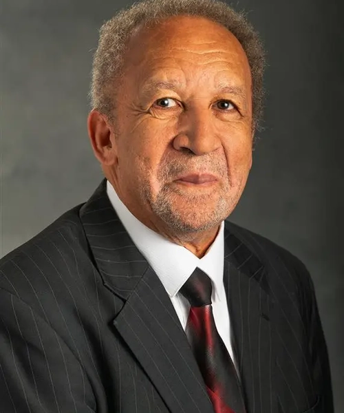 A photo of a man wearing a black suit, with a white button down underneath and a black and red multicolored tie against a dark gray background.