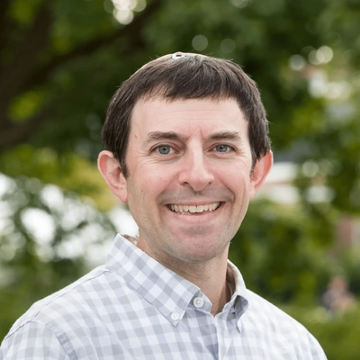 Portrait of a smiling person with short dark hair wearing a light blue checkered button-down shirt, standing outdoors with blurred green trees in the background.
