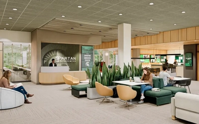 Interior view of the Spartan Success Center showing a reception desk, green seating areas, shared tables and indoor plants. People work on laptops throughout the space, and signage reading “Spartan Success Center” is visible on the back wall.