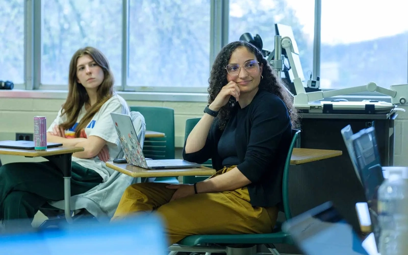 Two students listen while in a classroom, seated at their desks with laptops