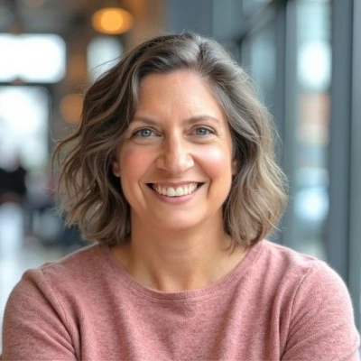 Smiling woman with shoulder-length wavy brown-and-gray hair wearing a pink sweater, seated indoors near large windows with a softly blurred background.