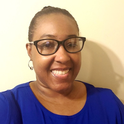 A person wearing a blue shirt and hoop earrings, standing against a plain light-colored wall.