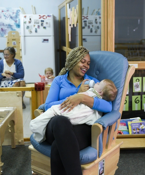 Woman sits in a chair feeding a bottle to a baby.