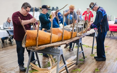 A group of participants shape a birch bark canoe around a wooden frame secured with upright stakes and concrete blocks.