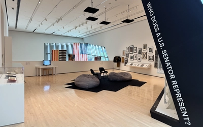 Interior view of an art gallery exhibit with large slanted text wall that says "Who does a U.S. Senator Represent?", beanbag seating artwork displays, U.S. flags and architectural windows.