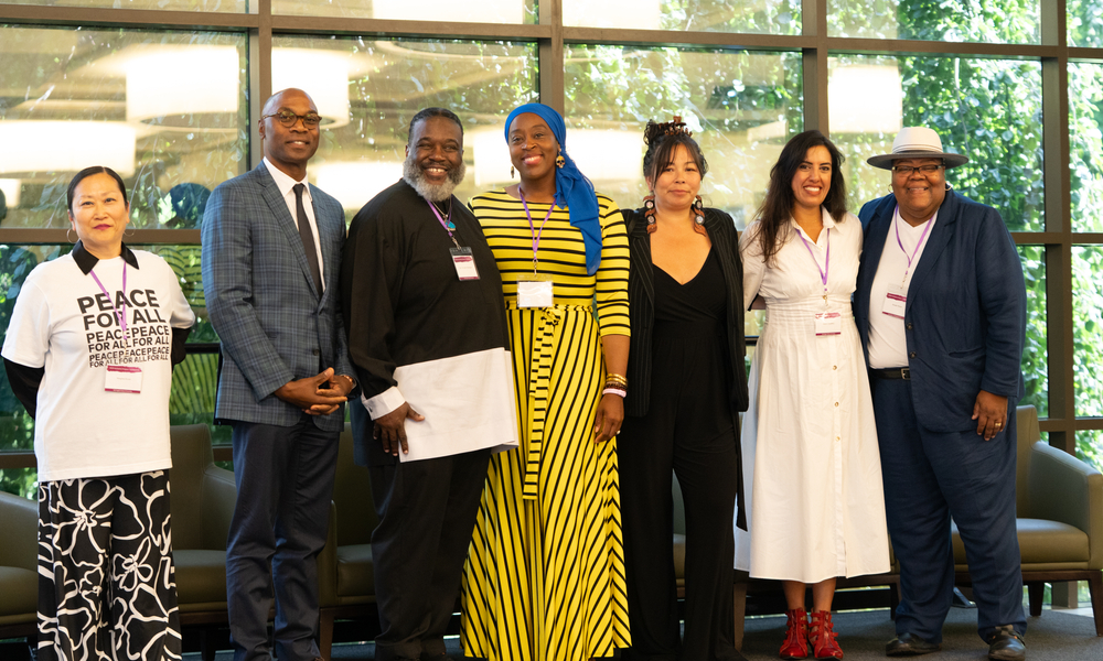A group of seven individuals stands side-by-side posing for a photo in front of a large grid window overlooking green trees. The group is dressed in a mix of professional attire and vibrant colors.