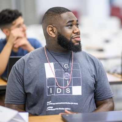 A close up of a young man sitting at a desk listening to a lecture. He is wearing a dark grey tishirt. He has a dark beard.