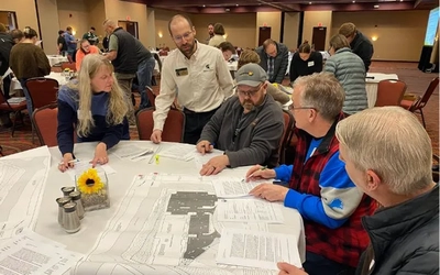 A group of people sit at a round table inspecting papers spread out atop the table