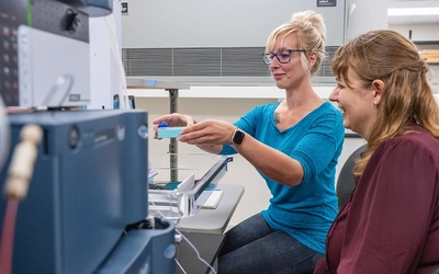 Two researchers handle samples beside a large research instrument.