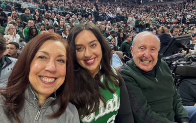 Three people seated courtside take a selfie at a packed basketball arena, with a large crowd wearing green visible in the stands behind them.