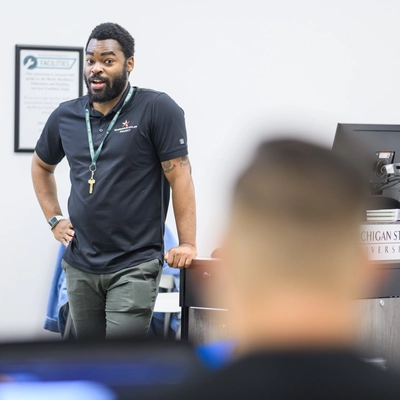 A man stands at a podium during a seminar. He is wearing a navy blue polo and army  green chinos