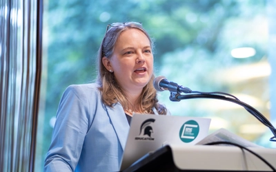 A speaker in a light blue blazer presents at a podium with a microphone and laptop during an education policy event, with large windows and greenery blurred in the background.