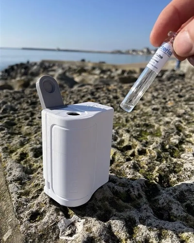 A Bactiquick test kit sitting on the sand at a beach as a hand reaches to insert a test tube into it.