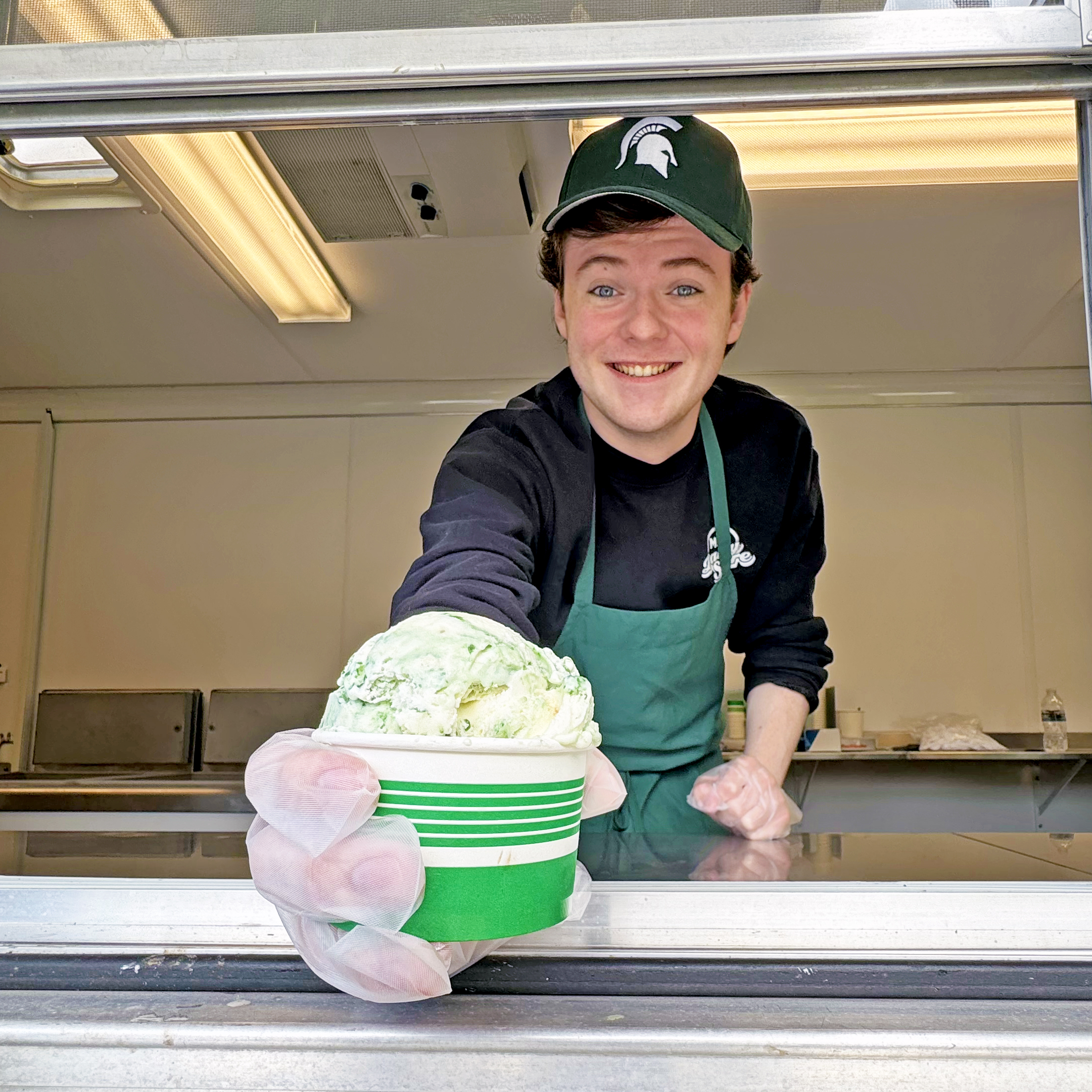 A smiling MSU Dairy Store employee leans out of the service window of the Dairy Store’s new ice cream trailer, holding out a cup of green-and-white ice cream. Wearing a green Spartan hat and apron, the employee serves customers from inside the mobile trailer on campus.