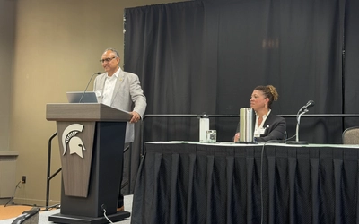 Two individuals stand at a podium and table to deliver remarks at a conference.