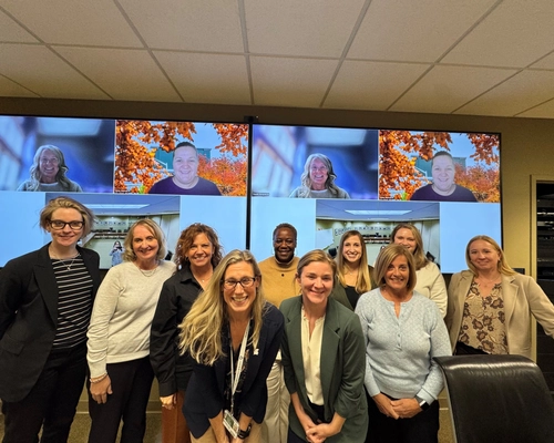 A group of ten people posing for a professional group photo in a conference room. They are standing in two rows, smiling at the camera. In the background, two large monitors display a video call with four additional remote participants and views of a secondary meeting space. The room has neutral-colored walls and a drop ceiling.