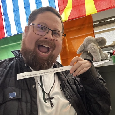 A man with an excited expression holds a slip of paper. An elephant plush and Mario figure are on a cabinet behind him.