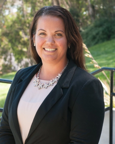 Professional headshot of Kim Pfotenhauer, DO, smiling in a black blazer and necklace with greenery in the background.
