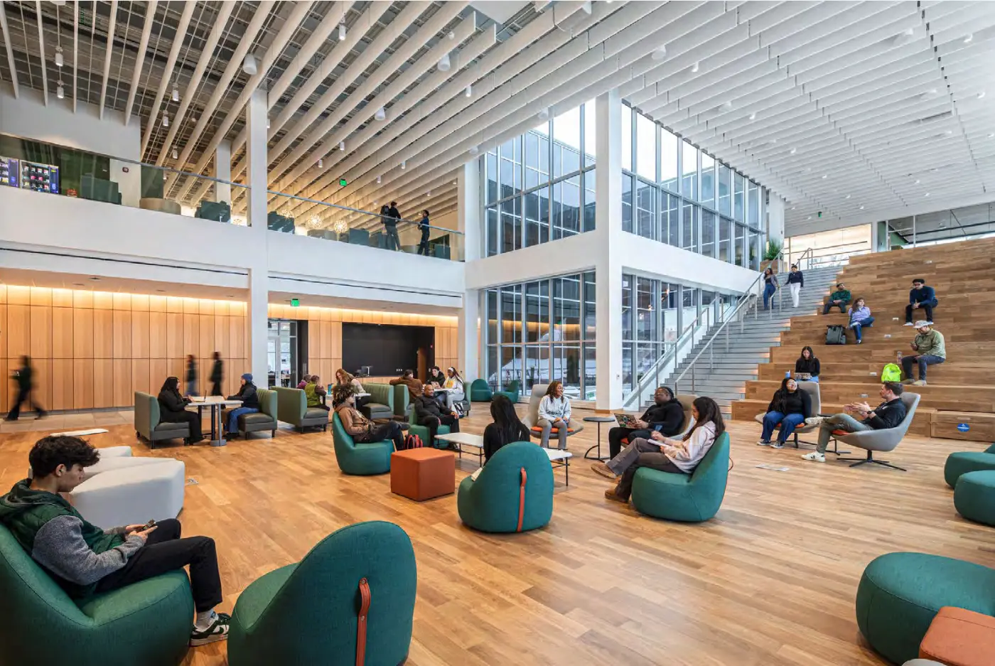 a two story open living room space inside a large campus building with flexible soft seating, an open wooden staircase, glass atrium and upstairs open balcony