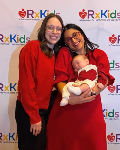 Rachel Hollander at an Rx Kids event, standing and smiling next to a woman and her baby.