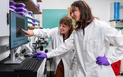 Two researchers in white lab coats, goggles, and purple gloves work at a microscope, gesturing at the instrument’s display.