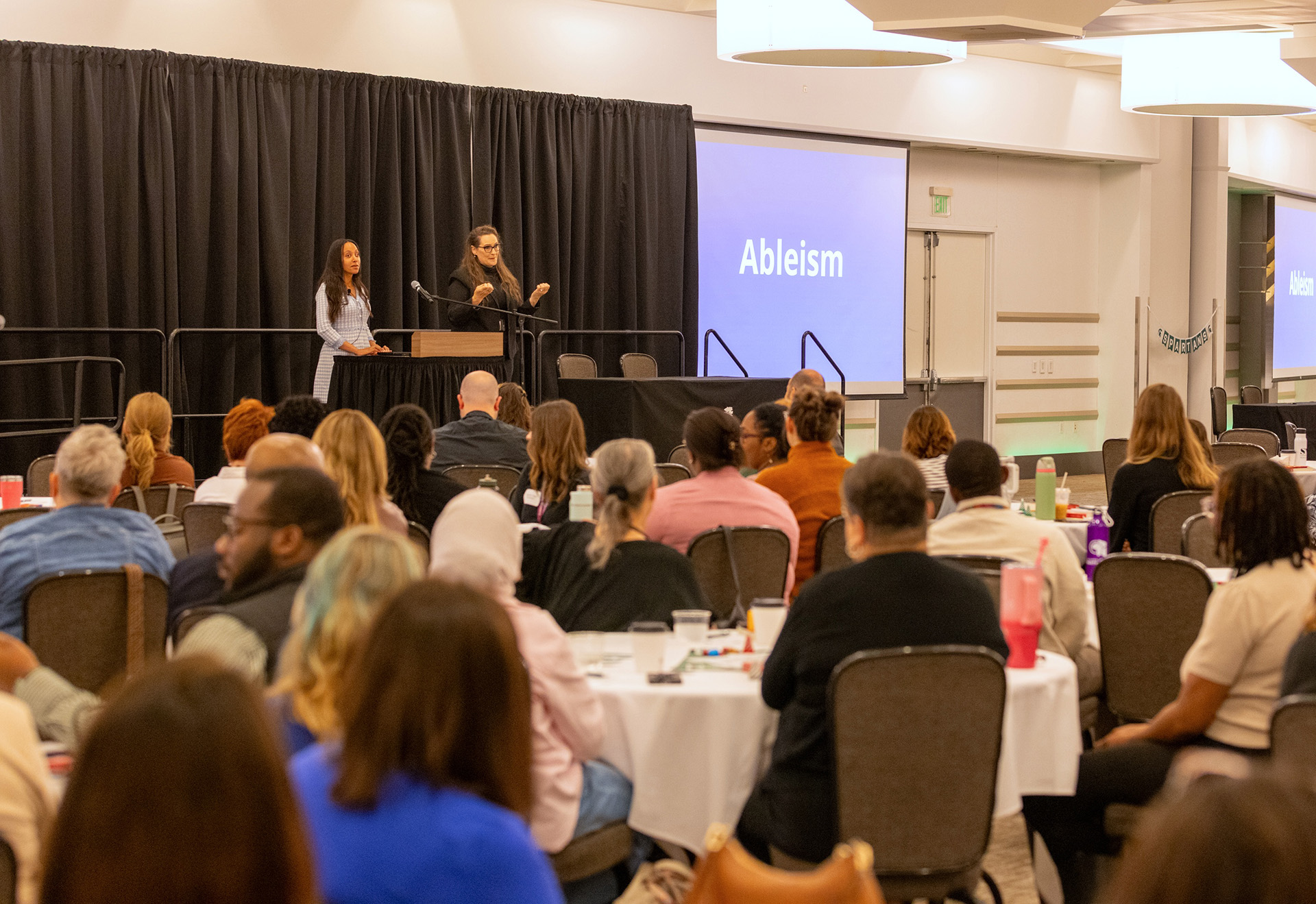 A speaker and an interpreter stand on a stage before a large audience, addressing them. The word Ableism is on a screen