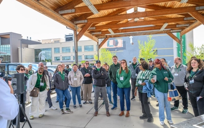 A group of people stand under a wooden pavilion listening to a presentation.