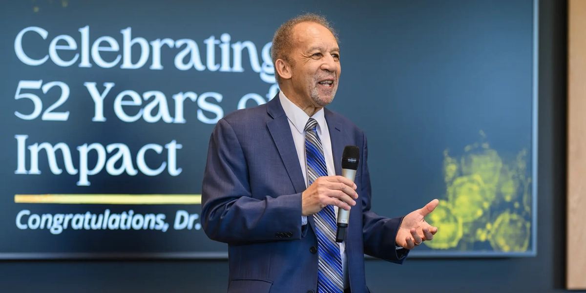 Photo of a man speaking in front of a screen that says "Celebrating 52 years of impact, Congratulations Dr. Lee June"
