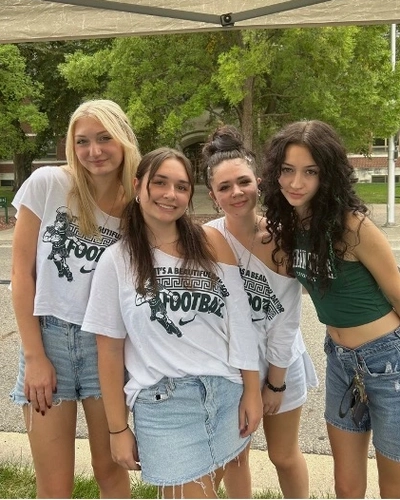 Four young women stand together smiling under a white pop-up canopy tent on campus.