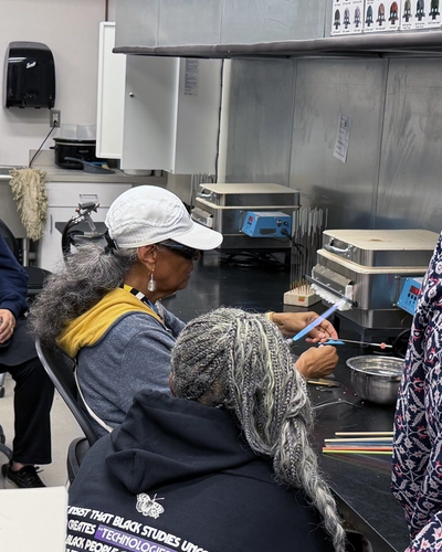 A participant wearing a navy SOLHOT 60+ hoodie watches as Dr. S.A. Simeon shapes glass over a flame at a beadmaking station.