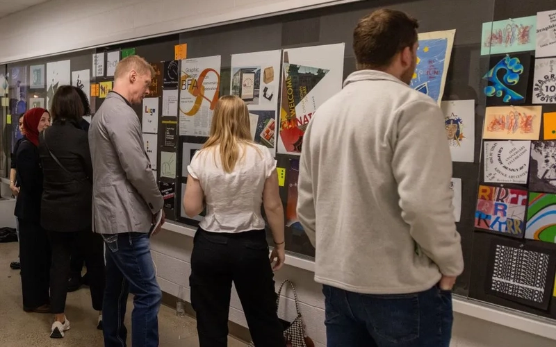 People line up to look at graphic design artwork posted behind glass on a black background.