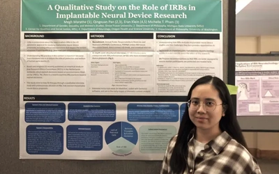 A young woman with long dark hair, wearing glasses and a brown-and-white plaid shirt, stands smiling slightly in front of a research poster. The poster's header reads "A Qualitative Study on the Role of IRBs in Implantable Neural Device Research" against a teal background.