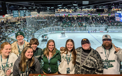 Seven individuals in MSU gear standing in a row. A crowded arena and ice rink are visible in the background.