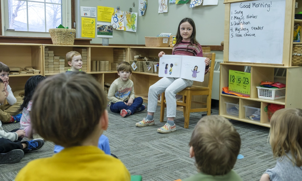 10-year-old sits on a chair reading a book to the surrounding children sitting on the floor.