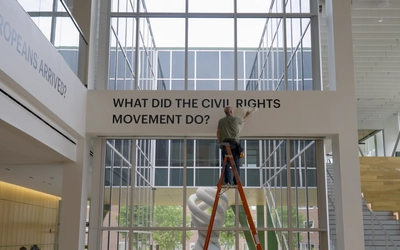 A person on a ladder installs letters for an exhibition that says "What did the Civil Rights Movement Do?" is put up along a wall of windows.
