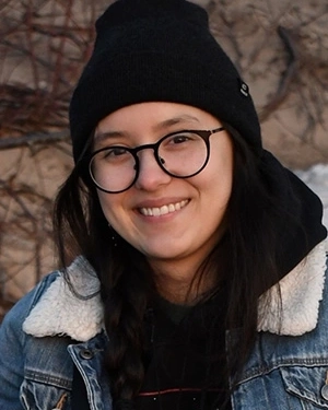 Close-up outdoor portrait of a smiling person wearing round black-rimmed glasses, a black beanie, a black hoodie under a denim jacket with a sherpa collar, and hair styled in a braid.