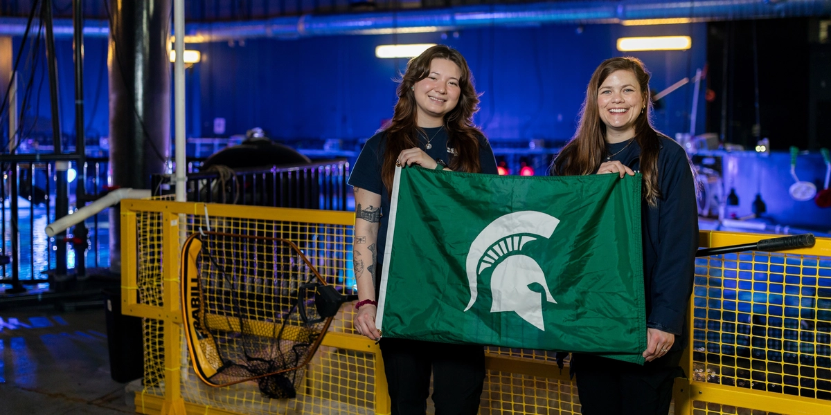 Dana MacCallumMhor and Lauren Grauer hold a Spartan helmet flag in front of yellow fencing at the Sea Life Michigan aquarium