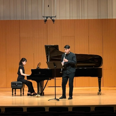A photo of elyor goforuv playing the clarinet. he is wearing a black suit and shirt and is standing on a stage in front of a grand piano.