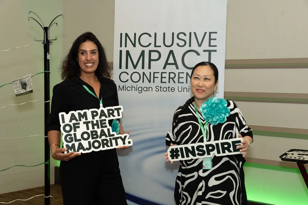 Two women hold photo prop signs in front of the Inclusive Impact Conference banner