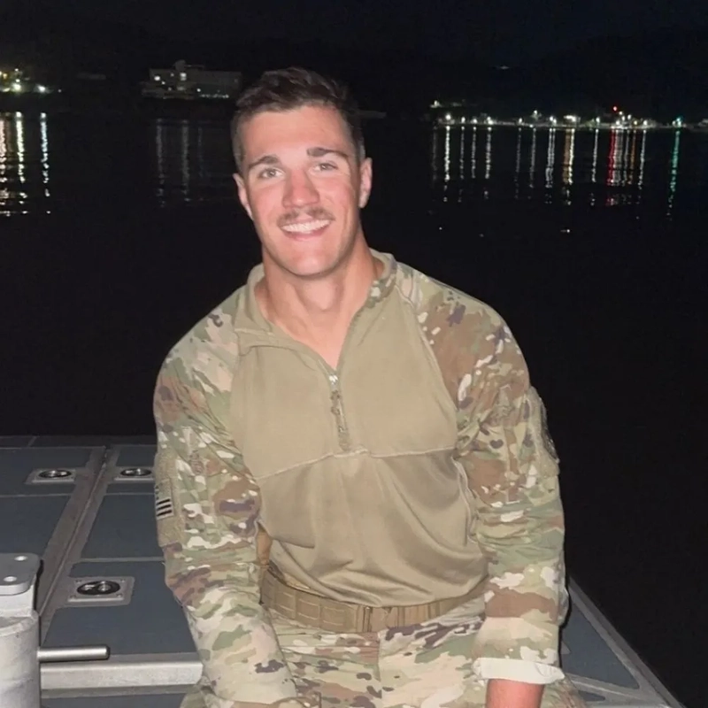 Student Colt Gray smiles outdoors at night in a military camouflage shirt with water and distant lights behind him.