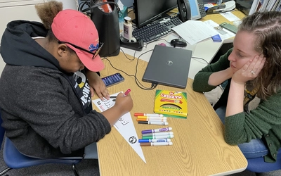 A high school student in a pink hat and black hoodie sits at a wooden desk, using a pink marker to color in a paper pennant. Seated across from her, a college adviser in a green cardigan rests her chin on her hand, observing the work. The desk is scattered with Crayola markers, a laptop, and a smartphone.