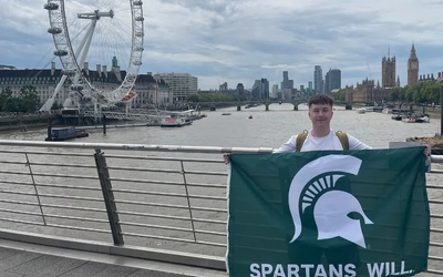 Liam McDonald holding up an MSU flag in front of the city of Cambridge while studying abroad at the University of Cambridge the summer after his freshman year.