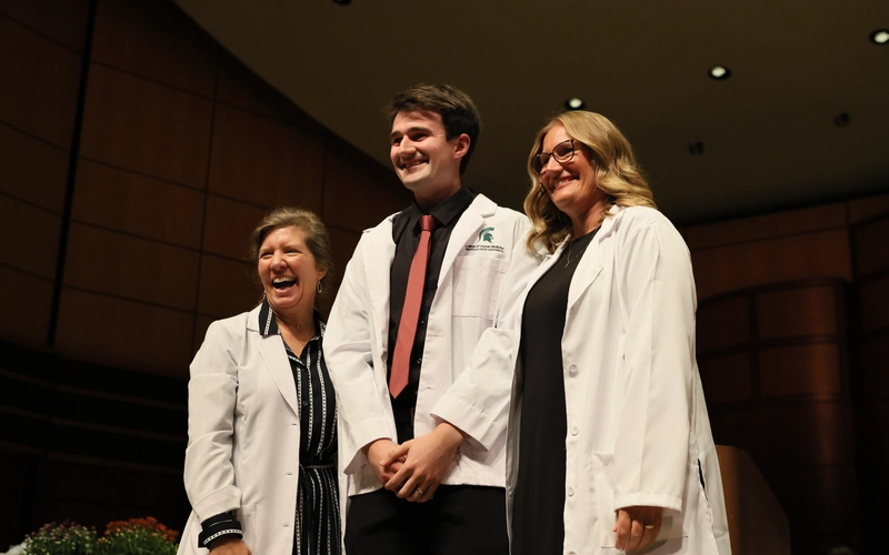 A medical student wearing a black button-down shirt, a dark pink tie, and a newly donned white medical coat with the Michigan State University College of Human Medicine logo patch on the left chest stands smiling on stage between two faculty members.