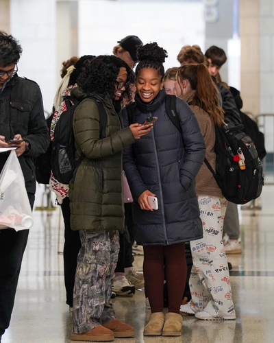 Two girls in winter coats laughing together while looking at a smartphone in a long indoor queue.