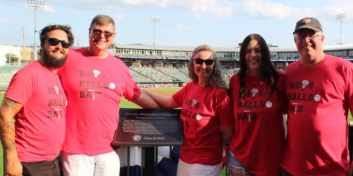 Group of people smile at the camera next to a plaque with a baseball field behind them.