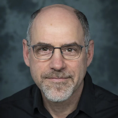 Studio headshot of a middle-aged man with a bald head, short gray facial hair, and glasses, wearing a black collared shirt against a blue-gray mottled background.