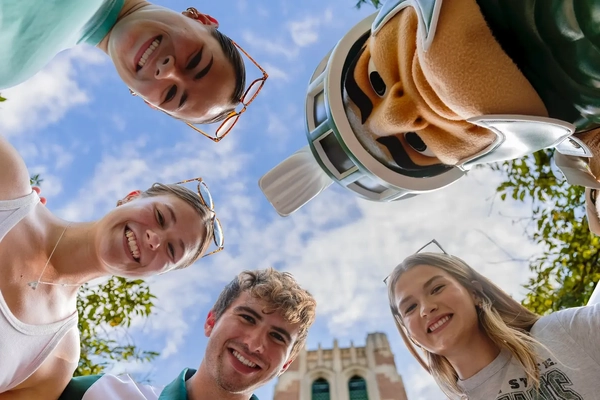 A group of smiling students lean in toward the camera outdoors on campus, joined by Sparty, with blue sky, trees and Beaumont Tower visible above them.