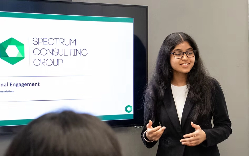 A person in a suit presents in an office setting with a computer screen next to them that reads SPECTRUM CONSULTING GROUP