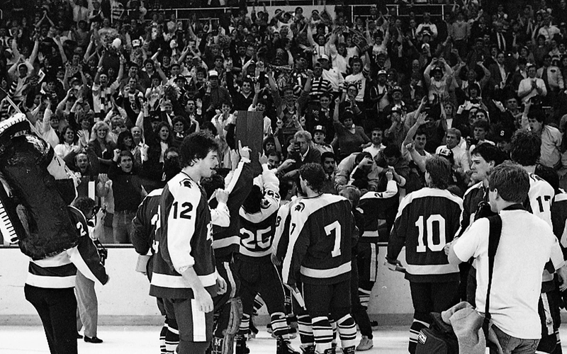 1986 MSU men's ice hockey team celebrating NCAA tournament win on the ice. In the background, fans can be seen cheering and a trophy is being lifted